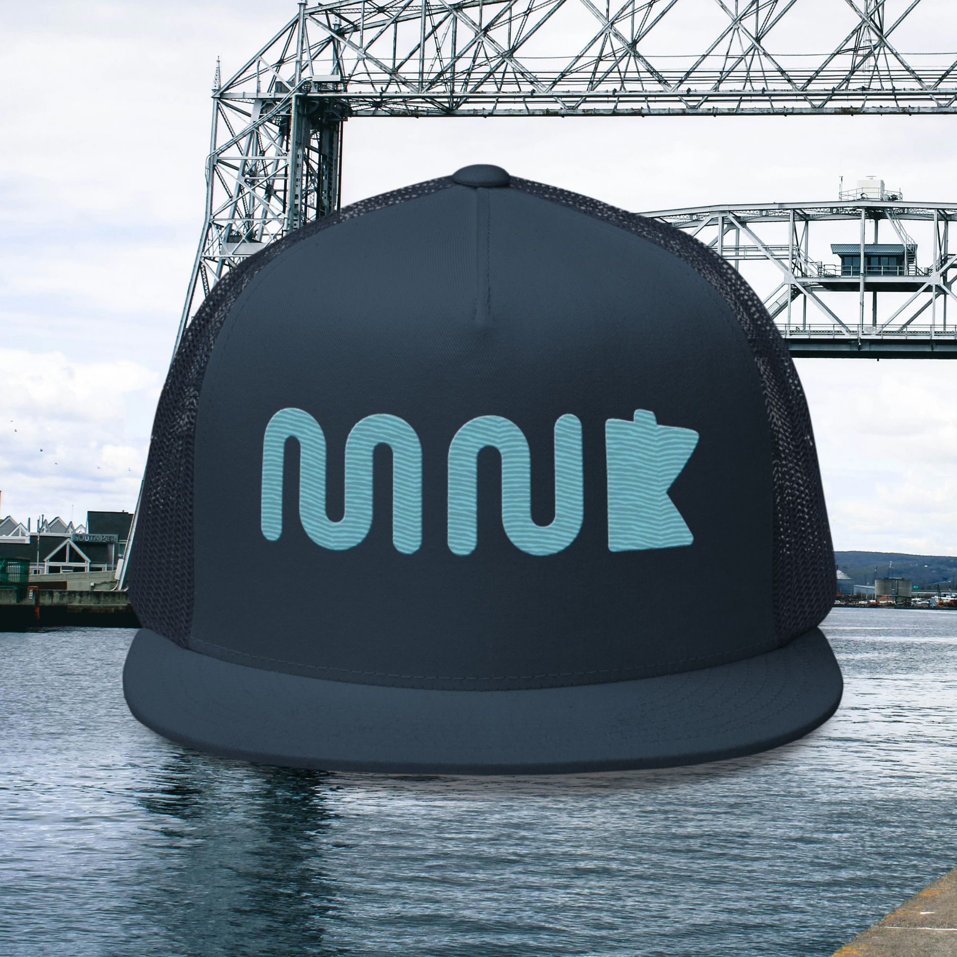 Minnesota hat with a blue "MN" using a wavy, river like font. There is a simplified blue outline of the state of Minnesota next to it. The midnight navy hat appears to be hovering over a photo of the aerial lift bridge in Duluth Minnesota.